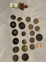 Top-down image showing 11 metal collectible buttons arranged with 9 pennies and a WW2 buddy brooch figurine on white background.