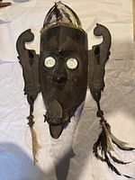 Front view of the large carved wooden mask with white circular eyes and feather adornments on the top and sides.