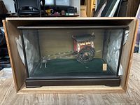 Full view of rickshaw model inside glass display case with black wooden frame and green felt base.