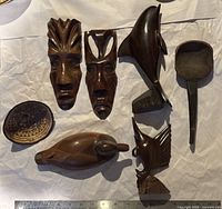 Seven wooden carvings arranged on white fabric including two masks, dolphin, ladle, bird, bird head, and round dish.