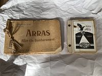Photo of two vintage books on a white crumpled paper background, the left book is a brown photo album titled 'ARRAS After the Bombardment 1914-1916', the right book is a smaller binder labeled 'Notes and Data'