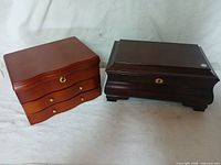 Two wooden jewelry boxes side by side on white background. The left is brown with three drawers, the right is black with a lock and curved feet.