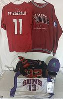 Photo showing multiple Arizona sports apparel items including Arizona Cardinals jersey #11 Fitzgerald, Cardinals pullover, Diamondbacks baseball cap, Dallas Cowboys Troy Aikman jersey, Phoenix Suns Charles Barkley #13 jersey, bobblehead, and collectible figurines.