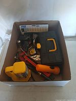 Box filled with various assorted hand tools visible from above including a black tool case, tape measure, yellow bottle jack, and orange screwdriver handle.