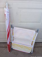 Photo showing the folded beach lounger with striped fabric in white, yellow, orange, and pink tones next to a folded beach umbrella wrapped in clear plastic with red and white stripes.