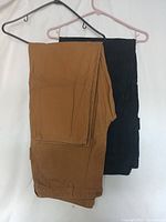 Two pairs of pants, one brown and one black, folded and hanging on hangers.