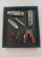 Top view of six camping multi-tool accessories in black display tray, showing variety of folding blades and pliers.