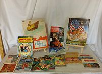 Photo of assorted children's books laid out on a white cloth showing a mix of storybooks and comics including titles like 'I Love America!', 'The Flying Trunk', and 'Popeye Goes on a Picnic'.