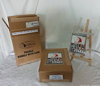 Two unopened brown cardboard boxes labeled 'clear acrylic frames w/ magnets' from AZAR Intl. Inc. One box is upright and the other is flat, both sealed and labeled with product info and quantity.
