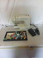 Off-white Kenmore sewing machine with attached black foot pedal and assorted sewing thread spools in front on a black tray.