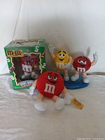 Four M&M collectible items: plush, two figures on display stand, and boxed candy dispenser.