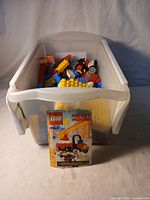 Plastic storage container filled with assorted Lego pieces and yellow base plates, with the original set 4164 instruction booklet propped in front.