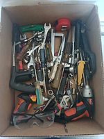 Top-down image of a cardboard box filled with assorted hand tools, two cordless drills, tape measure, and safety glasses visible on top.