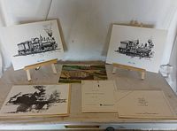 Five black and white vintage train prints arranged on a surface with two trackage maps placed in front. Two prints displayed on wooden easels. Prints show steam locomotives in detailed artistic style. Maps have a colorful design and depict train tracks.