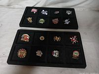 View of 15 assorted youth sports pins displayed in two separate black tray holders. Pins show a variety of youth sports themes with different team logos, cartoon mascots, and baseball equipment motifs.