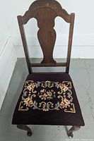 Photo showing front view of antique wooden chair with carved straight back and needlepoint upholstered seat cushion.