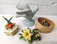 Top view of blue ceramic bird figurine, hexagonal hummingbird musical box, ceramic yellow lily flower with butterfly on wooden base, and small bird figurine in round box arranged against white wall.