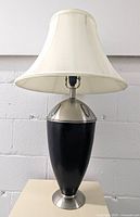 Full view of black and silver metal urn shaped table lamp with cream-colored lampshade showing the overall design and height.