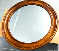 Front view of a large round mirror with a wooden frame laid flat on a table, showing the reflective glass and warm brown wood grain frame.