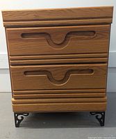 Photo of closed nightstand showing two drawers with carved cut-out handles, wood finish and black decorative metal legs.