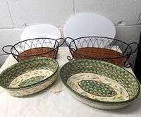 Two oval hand-painted ceramic casseroles with white plastic lids and wire and rattan carriers/trivets on a white lace tablecloth against a white wall.