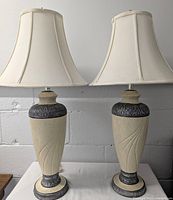 Pair of cream ceramic table lamps with pewter tone decorative bands and off-white fabric bell shades.