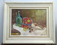 Framed oil painting showing a still life with a green glass bottle, a bowl of red and green apples, sliced bread, and nuts on a white cloth.