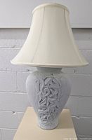 Full view of ceramic 3-way table lamp with ornamental leaf design on base and cream fabric lampshade, set against a white brick wall.