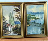 Two framed oil paintings side by side with gold-colored frames, one showing a church with two tall spires and a traditional half-timbered house with swans in the foreground; other a lake view with sailboats and a stone bridge.