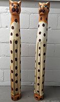 Front view of two tall wooden wild cat sculptures with elongated bodies and brown spots, standing against a white brick wall.