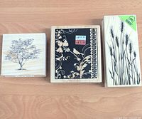 Top view of three wood block rubber stamps showing wood mounts with tree, floral border, and wheat stalk designs in black print