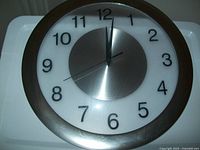 Front view showing 12-inch analog wall clock with white face, black numerals, silver central disc and rim, and clear plastic dome cover.