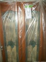 Pair of wooden window shutters with stained glass floral panels, covered in protective plastic wrap. Each pane shows blue flower with blue border on a white background inside oak frame, with knobs and hook closures visible.