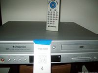 Front view of silver Polaroid VHS/DVD combo player with remote control standing on top, showing VHS and DVD slots, buttons, AV inputs and digital display.