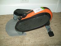 Side view of elliptical foot bike showing two large black foot pedals, orange casing, and tension dial