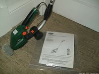 Green cordless garden shrub shear attached to silver extension pole, placed on floor with instruction manual beside it.