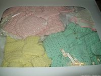 Photo showing seven hand-made crocheted infant outfits in pastel colors pink, green, yellow, and white, including dresses, cardigans, and booties.