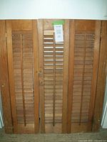 Front view of pair of stained wood louvre shutters, showing three vertical panels with horizontal slats and off-center hook closure.