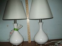 Two white ceramic bedroom lamps with acrylic bases and off-white shades with a ruler to show 18.5 inch height.