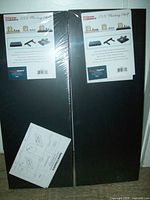 Pair of two black Home Basics floating shelves in plastic wrapping showing the front packaging label and product images