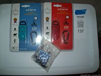 Front view of two personal portable emergency alarms in original blue and red packaging with visible metal clip accessories, and a third unpackaged blue alarm device with keychain, all placed on a white surface.