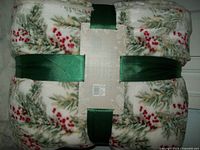 Front view of folded blanket with holiday branch and red berry pattern, tied with green ribbon and label in the middle.
