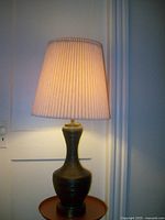 Table lamp lit on wooden surface, showing its ceramic jar body with ridged texture and pleated cream lampshade.