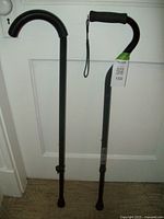 Photo showing two black adjustable walking canes, one with a curved handle and the other with a straight foam-covered handle and wrist strap, standing upright against a white door.