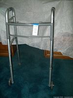 Front view of the silver metal folding walker showing two front wheels and two rear legs with rubber tips on a green carpet.