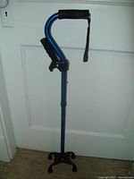 Full view of blue adjustable walking cane showing ergonomic black foam handle with attached wrist strap, side handle, and four rubber feet stand base.