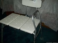 Front and side view of a white therapy or senior aid tub/shower bench with adjustable metal legs and a padded backrest, designed to span a bathtub for sitting and sliding into the tub safely.