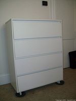 Full view of white melamine bureau standing on black round feet against white wall.