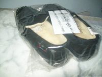 Pair of black Alpine Swiss moccasin slippers with cream fleece lining, packaged in clear plastic bag