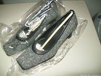 Top angled view of pair of black tweed pumps wrapped in clear plastic with white shoe shapers inside to maintain shape.